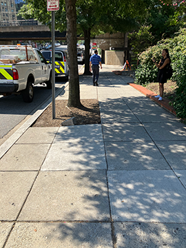 A picture of a sidewalk with a downward slope. To the right is the majority of the sidewalk along with a woman standing on the barrier for some landscaping. To the left is the street with several service trucks parked along the street edge. Directly ahead are a couple plots of dirt with trees that are casting shade on the sidewalk.